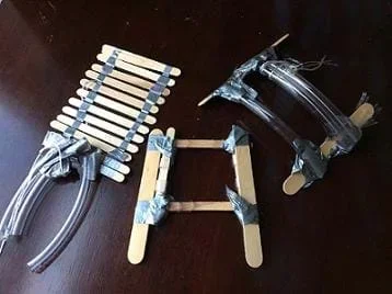 A photograph shows three different hand-device prototypes. All are made of duct tape and regular- and jumbo-sized craft sticks. Two of them also use clear plastic tubing and string.
