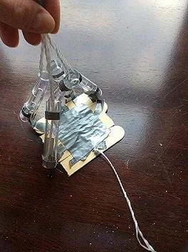 A photograph shows an assistive hand device prototype composed of four jumbo-sized craft sticks, cut in half and secured by duct tape. This piece is laid flat against a surface, representing the palm of a hand. Short, finger-like appendages, made by clear tubing pieces, are attached to the "palm' and are lifted by strings threaded through the tubular fingers.